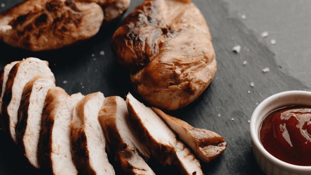 Sliced barbecued meat sits on a board next to a small dish of barbecue sauce